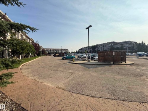 Asphalt parking area featuring multiple residential buildings in the background, mature landscaping, and a patterned concrete sidewalk - #6125 7331 South Terwillegar Dr Nw, Edmonton, AB - Outdoor With View