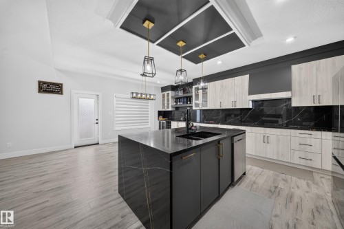 Kitchen featuring a large island with an integrated sink, contrasting upper and lower cabinetry, black marble-look countertops and backsplash, and light wood-finish flooring - 3217 Pelerin Crescent, Beaumont, AB - Indoor Photo Showing Kitchen With Double Sink With Upgraded Kitchen