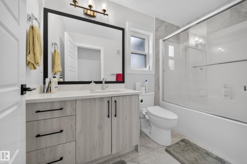 Bathroom featuring a single vanity with stone countertop, wood-finish cabinetry, a framed mirror, and a shower-tub combination with a glass enclosure - 3217 Pelerin Crescent, Beaumont, AB - Indoor Photo Showing Bathroom
