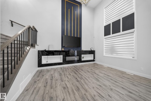 Wood-finish flooring, two-story window with horizontal blinds, built-in media console with a black marble-look finish, staircase with carpeted treads and metal balusters, and a decorative wall panel - 3217 Pelerin Crescent, Beaumont, AB - Indoor Photo Showing Other Room