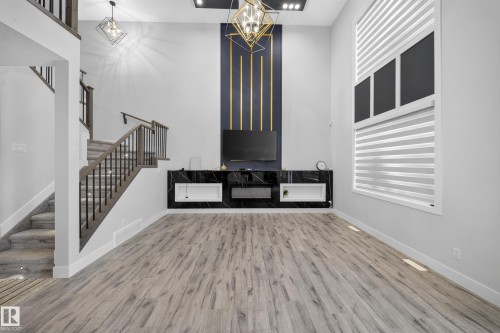 Two-story ceiling, wood-finish flooring, built-in media console, architectural wall accent with gold-tone detailing, and a multi-level staircase with metal balusters - 3217 Pelerin Crescent, Beaumont, AB - Indoor Photo Showing Other Room