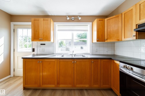 Kitchen featuring wood-finish cabinetry, a double basin stainless steel sink, black countertops, white square tile backsplash, and wood-finish flooring - 115 Griesbach Road Nw, Edmonton, AB - Indoor Photo Showing Kitchen