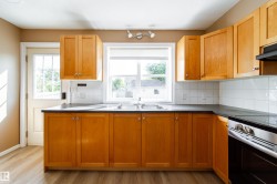 Kitchen featuring wood-finish cabinetry, a double basin stainless steel sink, black countertops, white square tile backsplash, and wood-finish flooring - 