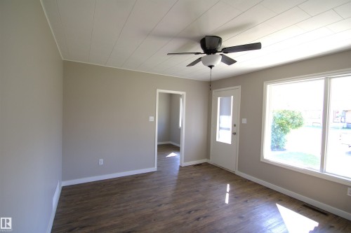 Wood-finish flooring throughout, neutral wall tones, white trim, ceiling fan with light fixture, and large windows - 4706 51 Ave, St. Paul Town, AB - Indoor Photo Showing Other Room