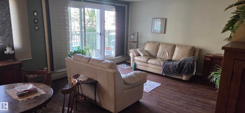 Wood-finish flooring throughout, complemented by light-colored walls and a sliding glass door with balcony access - #215 4404 122 St Nw, Edmonton, AB - Indoor Photo Showing Living Room