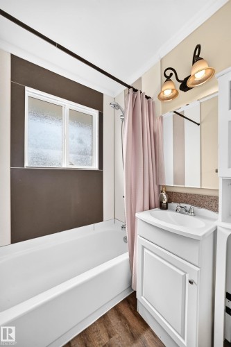White bathtub with shower, white vanity cabinet with sink, decorative wall sconce lighting, and wood-finish flooring - 50 Rocque, Fort Saskatchewan, AB - Indoor Photo Showing Bathroom