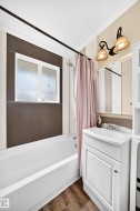 White bathtub with shower, white vanity cabinet with sink, decorative wall sconce lighting, and wood-finish flooring - 