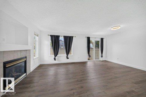 Spacious room featuring wood-finish flooring, a tile-surround fireplace, multiple windows, a sliding glass door, and an overhead flush-mount light fixture - #47 30 Levasseur Rd, St. Albert, AB - Indoor Photo Showing Living Room With Fireplace
