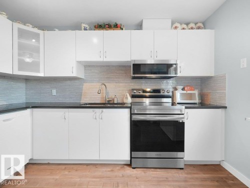 White cabinetry, dark countertops, a tile backsplash, stainless steel appliances, and wood-finish flooring - #54 2131 Oak St, Sherwood Park, AB - Indoor Photo Showing Kitchen With Upgraded Kitchen