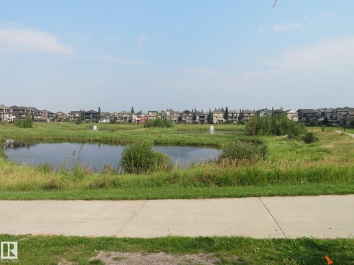 Paved walking path bordering a tranquil pond with aeration fountains, surrounded by natural landscaping and residential architecture - 3233 Winspear Crescent Sw, Edmonton, AB - Outdoor With View
