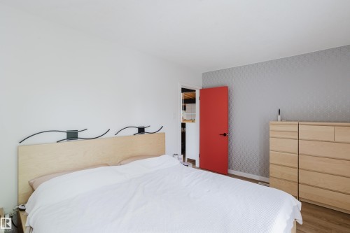 Wood-finish flooring with a light wood headboard, contrasting red door, and a patterned accent wall - 9059 52 Street Nw, Edmonton, AB - Indoor Photo Showing Bedroom