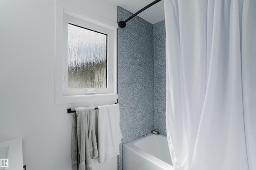 White bathtub with a curved shower rod, patterned blue-gray wall tile, and a privacy window - 9059 52 Street Nw, Edmonton, AB - Indoor Photo Showing Bathroom