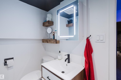 Illuminated vanity mirror, floating wood shelves, wood-finish vanity with integrated sink, black faucet, and white walls - 9059 52 Street Nw, Edmonton, AB - Indoor Photo Showing Bathroom