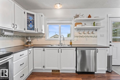 White cabinetry with black hardware, dark countertops, a stainless steel dishwasher, a double basin sink with a high-arc faucet, and wood-finish flooring - 5330 43 Street, Cold Lake, AB - Indoor Photo Showing Kitchen With Double Sink