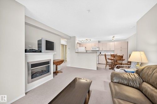 Integrated living area with a built-in fireplace, neutral carpeting, and a kitchen featuring light cabinetry, a tile backsplash, and stainless-steel appliances - #216 17003 67 Av Nw, Edmonton, AB - Indoor Photo Showing Living Room With Fireplace