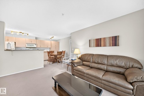Combined living and dining areas featuring light-toned carpeting, a partial wall separating the kitchen, light wood-finish cabinetry, and track lighting fixtures - #216 17003 67 Av Nw, Edmonton, AB - Indoor Photo Showing Living Room