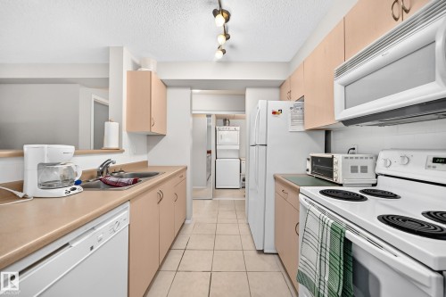 Galley kitchen featuring light wood-finish cabinetry, solid surface countertops, a stainless steel sink, and white appliances - #216 17003 67 Av Nw, Edmonton, AB - Indoor Photo Showing Kitchen With Double Sink