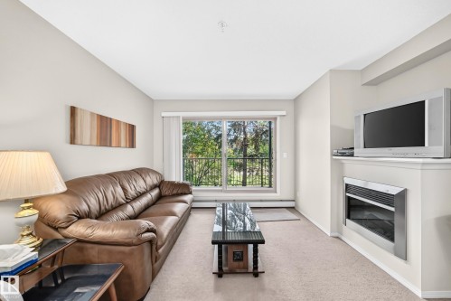 Neutral wall paint, built-in electric fireplace, sliding glass door, balcony with iron railing, and carpeted flooring - #216 17003 67 Av Nw, Edmonton, AB - Indoor Photo Showing Living Room With Fireplace