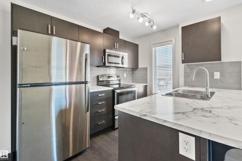 Stainless steel appliances, dark wood-finish cabinetry, light marble-look countertops, and a gray subway tile backsplash - #318 5521 7 Av Sw Sw, Edmonton, AB - Indoor Photo Showing Kitchen With Stainless Steel Kitchen With Double Sink With Upgraded Kitchen