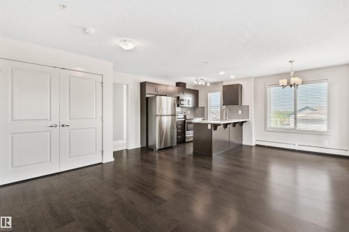 Open-concept living area with dark wood-finish flooring, white walls, and a kitchen featuring stainless steel appliances, dark cabinetry, and an island with bar seating - #318 5521 7 Av Sw Sw, Edmonton, AB - Indoor Photo Showing Kitchen