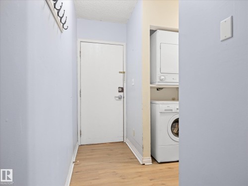 Wood-finish flooring complementing a built-in coat rack, a white door with a deadbolt, and a stacked washer and dryer unit - #110 10139 117 St Nw, Edmonton, AB - Indoor Photo Showing Laundry Room