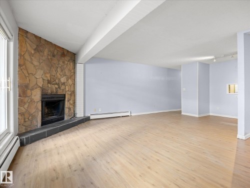 Spacious room featuring wood-finish flooring, a stone-clad fireplace with a black hearth, and track lighting - #110 10139 117 St Nw, Edmonton, AB - Indoor With Fireplace