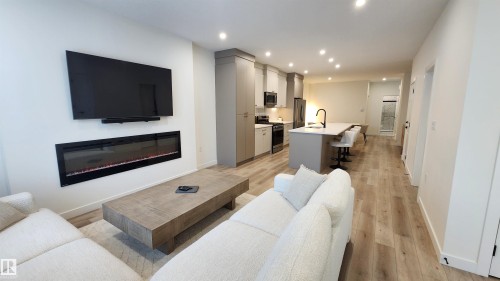 Open-concept living space with wood-finish flooring, a contemporary electric fireplace, a kitchen island with seating, recessed lighting, and modern cabinetry - 220 28 Street Sw, Edmonton, AB - Indoor Photo Showing Living Room With Fireplace