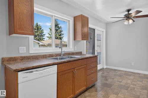 Wood-finish cabinetry, stainless steel double basin sink, white dishwasher, ceiling fan with integrated lighting, and patterned flooring - 13214 123 Street Nw, Edmonton, AB 