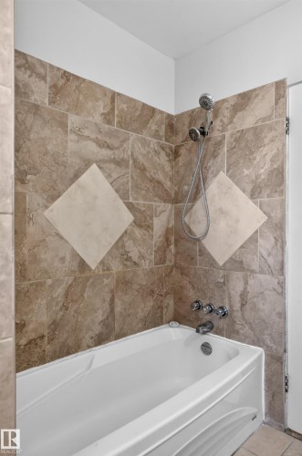 White bathtub with polished chrome fixtures, surrounded by neutral-toned tiled walls featuring a decorative diamond pattern - 13214 123 Street Nw, Edmonton, AB 