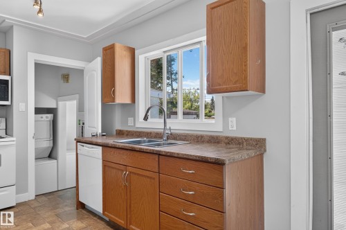 Wood-finish cabinetry, built-in dishwasher, stainless steel sink with gooseneck faucet, window with white trim, and a stacked washer and dryer in an adjacent alcove - 13214 123 Street Nw, Edmonton, AB 