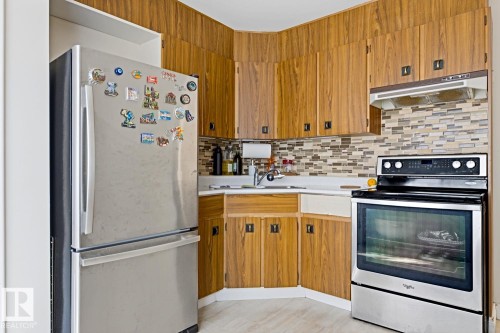 Efficient galley kitchen featuring warm wood cabinetry, stainless steel appliances including a gas range and built-in oven, mosaic tile backsplash, and a two-tier countertop layout with ample storage - 9740 63 Avenue Nw, Edmonton, AB 