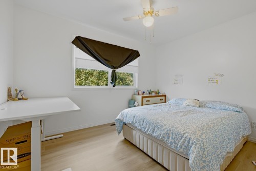 Bright secondary bedroom featuring wood-finish flooring, a large operable window with motorized blackout shade, white painted walls, and built-in desk workspace with integrated storage - 9740 63 Avenue Nw, Edmonton, AB 
