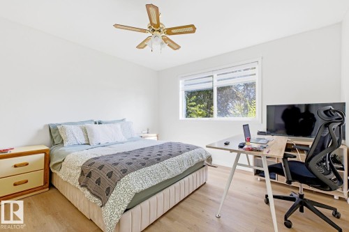 Bright bedroom with wood-finish flooring, large dual-pane windows with natural light, ceiling fan with brass finish, wood nightstand with drawer storage, and dedicated workspace with desk and ergonomic seating - 9740 63 Avenue Nw, Edmonton, AB 
