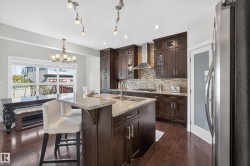 Kitchen featuring dark wood cabinetry, a granite-topped island with dual sinks, stainless steel appliances, a textured tile backsplash, and dark wood-finish flooring - 