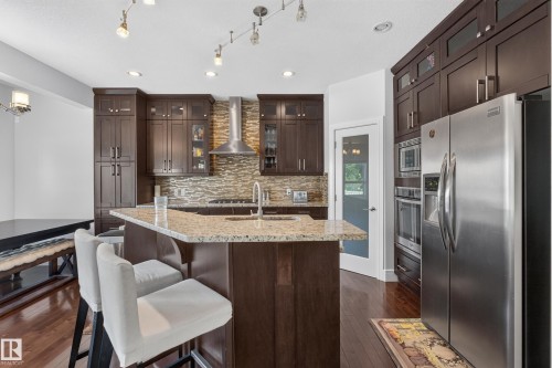 Kitchen featuring wood-finish flooring, dark wood cabinetry, stainless steel appliances, a stone-finish backsplash, and a large island with a light-toned countertop - 3685 Claxton Place Sw, Edmonton, AB 