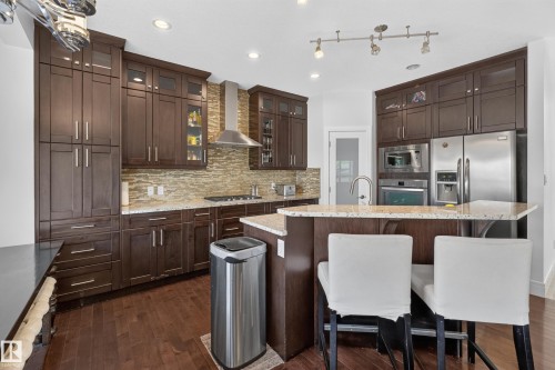 Kitchen featuring wood-finish flooring, dark wood cabinetry, light-toned countertops, a textured backsplash, and stainless steel appliances - 3685 Claxton Place Sw, Edmonton, AB 