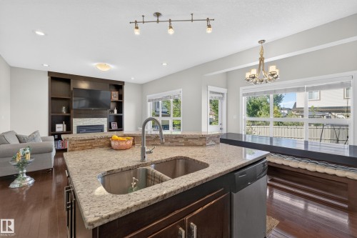 Kitchen island with a double basin sink and a high-arc faucet, granite-style countertops, wood-finish flooring, built-in entertainment center with shelving and a fireplace, and a dining area with a chandelier - 3685 Claxton Place Sw, Edmonton, AB 