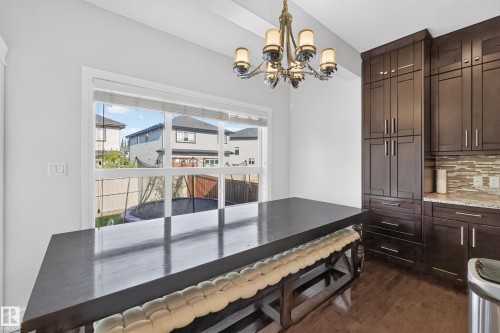 Dining area featuring an elegant chandelier, a large window, rich wood-finish flooring, and built-in dark wood cabinetry with a light-toned backsplash - 3685 Claxton Place Sw, Edmonton, AB 