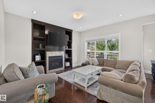 Dark wood-finish flooring, built-in shelving, stone-clad fireplace, recessed lighting, and a large window - 3685 Claxton Place Sw, Edmonton, AB 