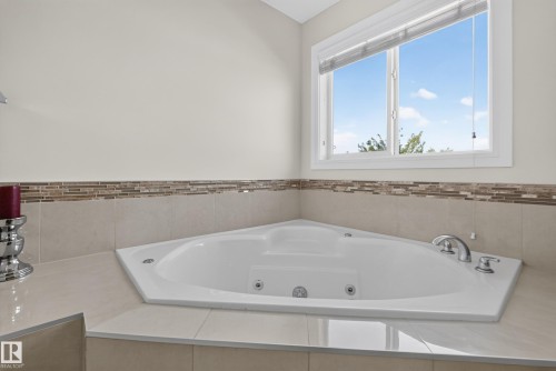 Corner jetted tub with polished chrome fixtures, surrounded by light-toned tile, with a decorative mosaic tile accent - 3685 Claxton Place Sw, Edmonton, AB 