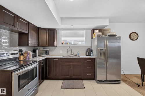 Kitchen featuring dark wood-finish cabinetry, light-colored countertops, stainless steel appliances, and a tiled backsplash - 3685 Claxton Place Sw, Edmonton, AB 