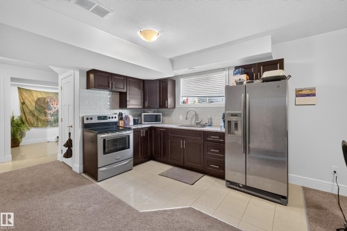 Compact kitchen featuring dark wood-finish cabinetry, stainless steel appliances, a white tile backsplash, and light-toned countertop surfaces - 3685 Claxton Place Sw, Edmonton, AB 