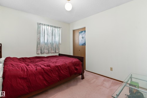 Spacious bedroom featuring hardwood-finish flooring, a wood-frame bed with deep burgundy bedding, sheer curtains at the window, and a solid wood entry door - 17724 94 Street Nw, Edmonton, AB 