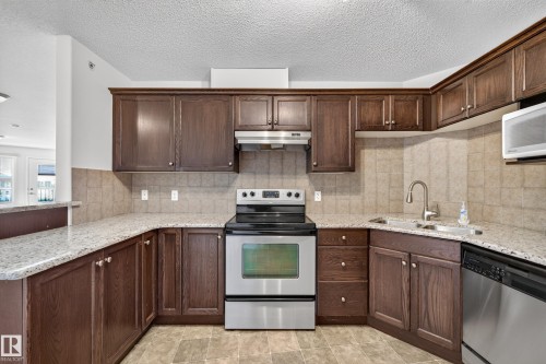 Kitchen featuring wood cabinetry, stainless steel appliances, speckled countertops, and a tiled backsplash - #6430 7331 South Terwillegar Dr Nw, Edmonton, AB 