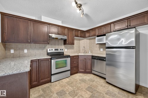 Kitchen featuring dark wood-finish cabinetry, light-toned stone-finish countertops, stainless steel appliances, and a tiled backsplash - #6430 7331 South Terwillegar Dr Nw, Edmonton, AB 