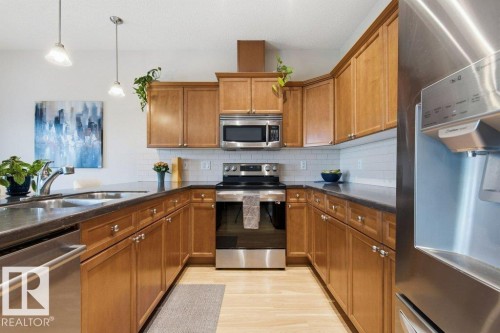 U-shaped kitchen featuring light wood cabinetry, dark countertops, white subway tile backsplash, stainless steel appliances, and wood-finish flooring - #17 7293 South Terwillegar Dr Nw, Edmonton, AB - Indoor Photo Showing Kitchen With Double Sink