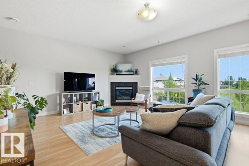 Spacious living area featuring wood-finish flooring, a corner fireplace with a dark tile surround and white mantel, and large windows - #17 7293 South Terwillegar Dr Nw, Edmonton, AB - Indoor Photo Showing Living Room With Fireplace