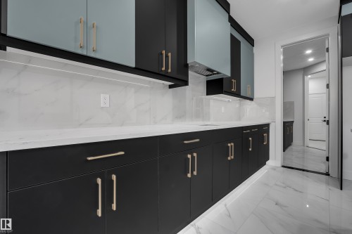 Contemporary kitchen featuring dark flat-panel cabinetry with gold-tone hardware, white marble-patterned countertops, a matching backsplash, and integrated under-cabinet lighting - 4005 39 Street, Beaumont, AB - Indoor
