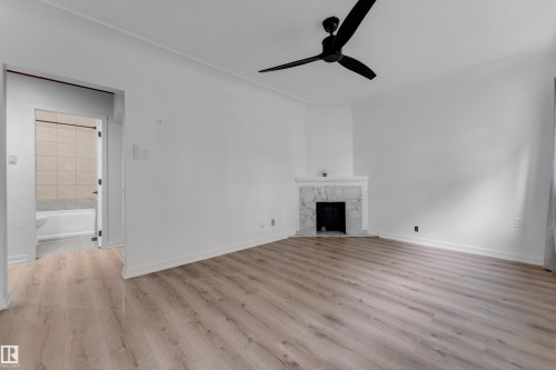Wood-finish flooring, white walls, black ceiling fan, white fireplace with marble surround, and an adjacent bathroom with a white tub and tiled walls - 12116 81 Street Nw, Edmonton, AB 