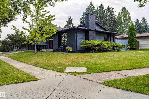 Contemporary home featuring a modern black exterior with geometric architectural lines, expansive manicured lawn with mature landscaping, concrete driveway with clean sightlines, and abundant natural tree coverage throughout the property - 14 Brunswick Crescent, St. Albert, AB 